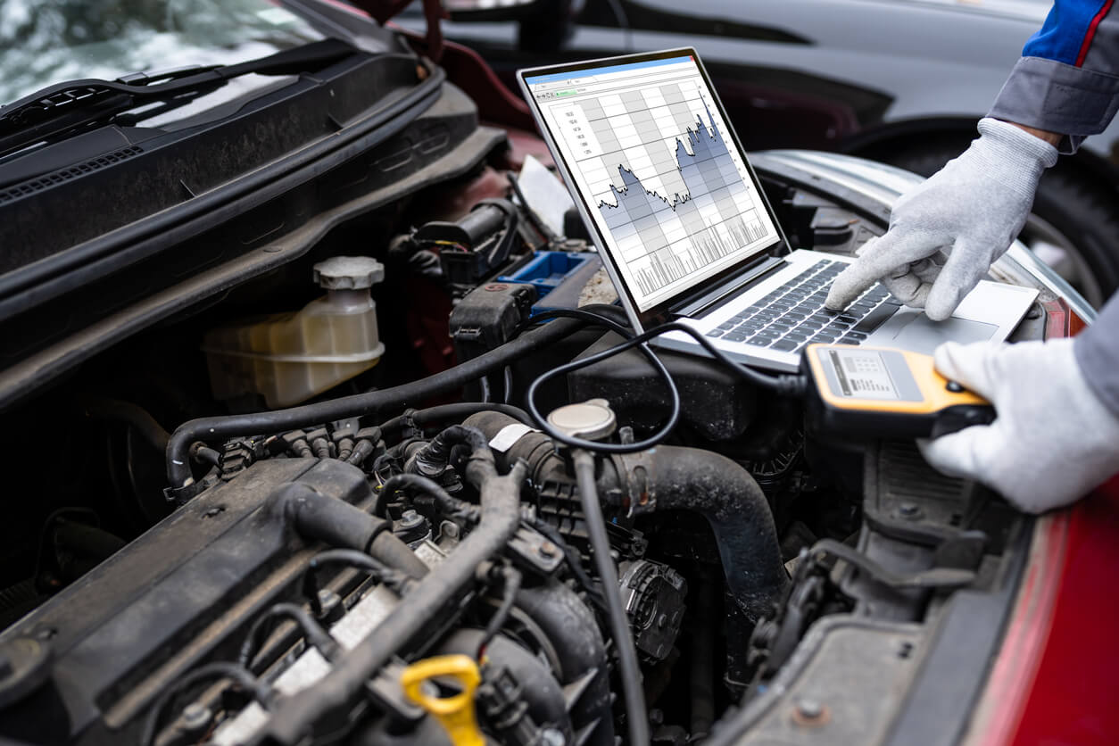Laptop open on top of a vehicle's engine, connected to the engine by cables, with laptop screen showing diagnostic information presented as a graph.